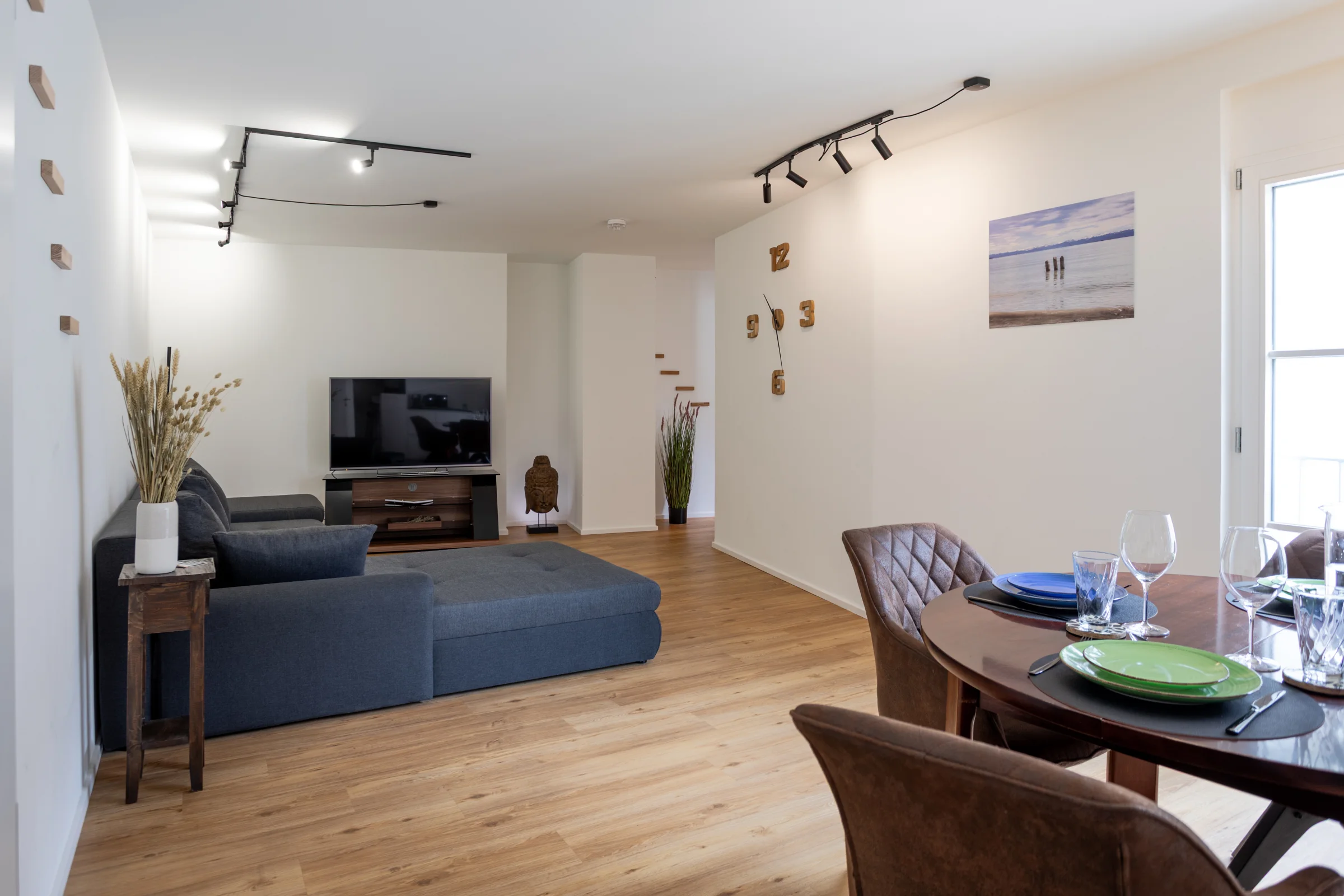 Heller Wohnbereich mit grauem Ecksofa, TV-Stand, Holzboden, Wand-Uhr aus Holzzahlen und gedecktem Esstisch