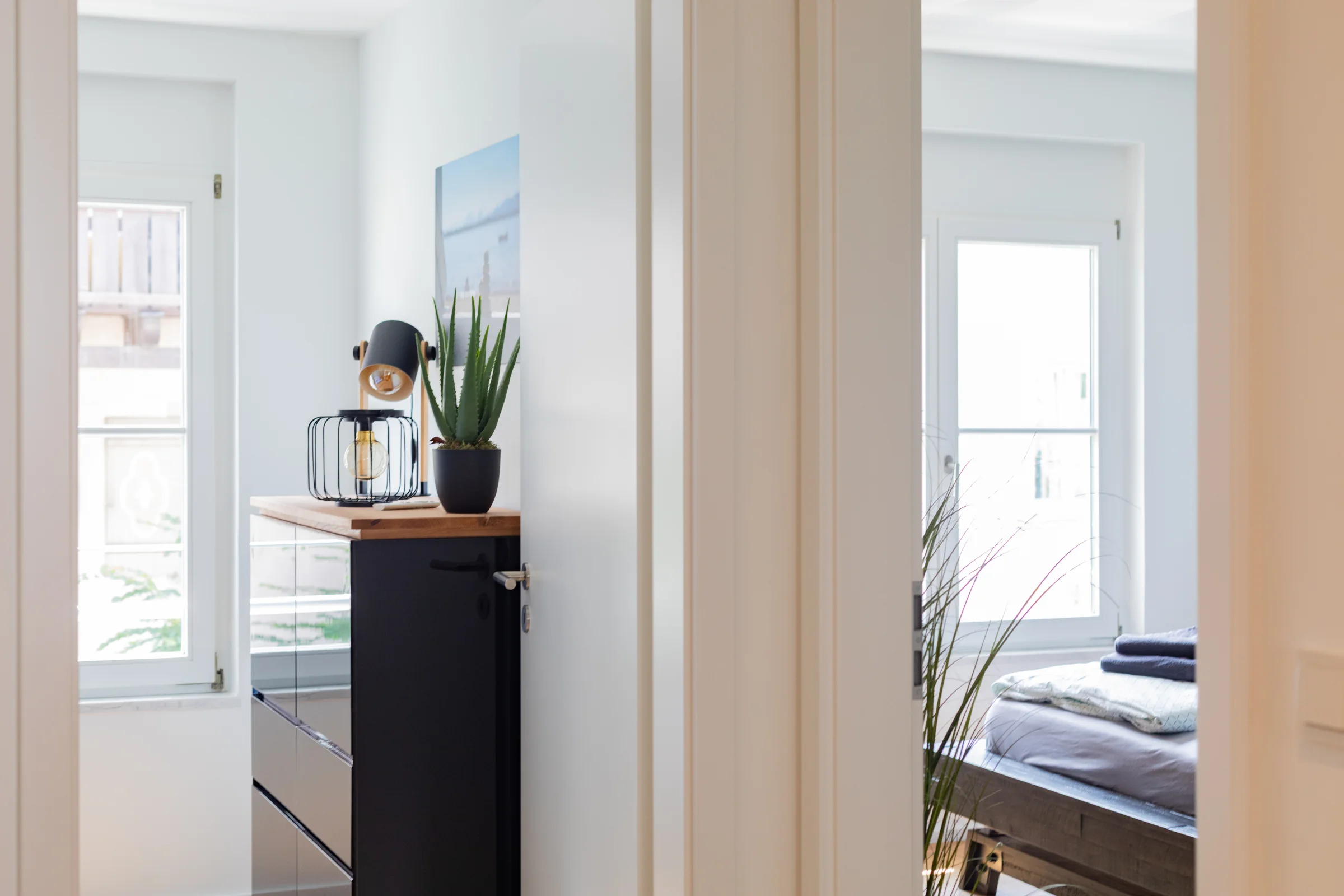 Schmale Kommode mit Drahtlampe und Topfpflanze, Blick durch offene Tür zum Fenster